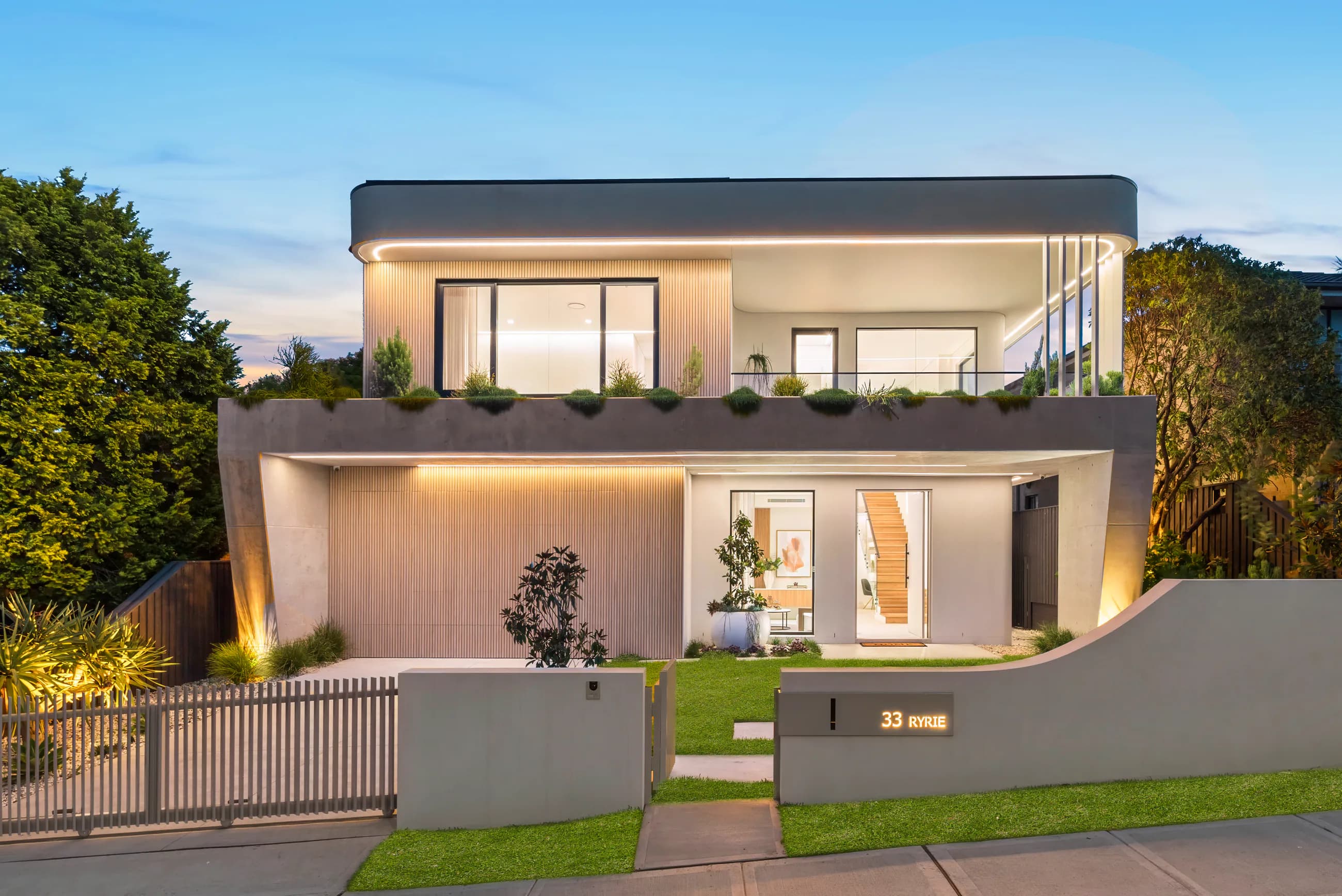Ryrie Residence street elevation with curved concrete forms, warm architectural lighting, and layered landscaping